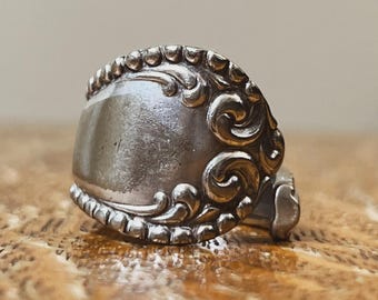 Op de afbeelding: Een zilveren ring met een sierlijk, vintage design. De ring heeft een rechthoekige voorkant met gedetailleerde krullen en een kralenrand. Het metaal heeft een licht aangeslagen uiterlijk, wat duidt op ouderdom en slijtage. De ring is waarschijnlijk een uniek, handgemaakt stuk.