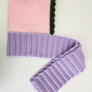 May include: A handmade crochet hood and scarf set in soft pastel colors. The hood is light pink with a dark green scalloped edge, transitioning to a lavender scarf. The set is made of a ribbed crochet stitch.