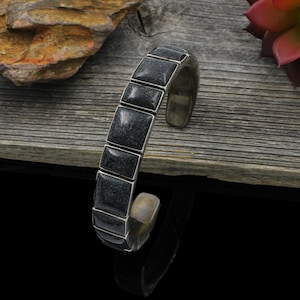 Pode incluir: Uma pulseira tipo bracelete com uma série de incrustações retangulares de pedra cinza-escura texturizada, inseridas numa armação de metal polido em tom prateado. A pulseira tem um design aberto, interior metálico liso e estrutura rígida.