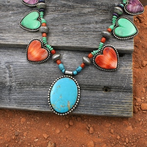 Puede incluir: Un collar turquesa y plata con colgantes en forma de corazón y ovalados. El collar presenta un gran colgante ovalado turquesa, colgantes en forma de corazón en tonos rojos, verdes y morados, y cuentas plateadas. El collar se exhibe sobre una superficie de madera envejecida.