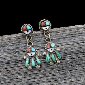 May include: A pair of silver-toned dangle earrings. Each earring features a circular top with a red, turquoise, and white inlay, connected by a chain to a figure with turquoise teardrop-shaped accents. The earrings are set against a weathered wooden surface.