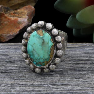 May include: A silver ring featuring an oval turquoise gemstone. The turquoise is a vivid blue-green with brown markings. The ring is encircled by small silver beads. The ring rests on a weathered wooden surface.