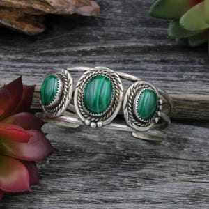 May include: A silver cuff bracelet with three oval malachite stones. Each stone is set within a decorative, twisted rope-like border. The bracelet has a vintage, handcrafted appearance, with a focus on natural materials and design.