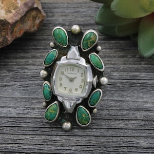 May include: A silver-toned Caravelle watch ring with a white face and turquoise accents. The watch has a rectangular face with silver hands and hour markers. The ring is surrounded by oval-shaped turquoise stones and silver beads.