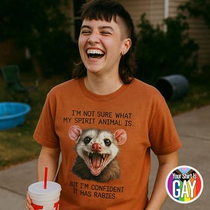 Peut inclure: T-shirt rouille avec un dessin d'opossum et le texte "I'M NOT SURE WHAT MY SPIRIT ANIMAL IS... BUT I'M CONFIDENT IT HAS RABIES." Un logo arc-en-ciel avec l'inscription "Your Shirt Is GAY" est en bas à droite.
