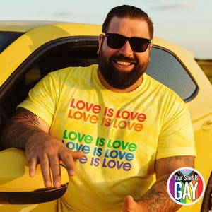 Può includere: Una maglietta gialla con la scritta "Love is Love" a colori arcobaleno. La persona che indossa la maglietta è appoggiata a un'auto gialla. È visibile un logo circolare con la scritta "Your Shirt Is GAY".