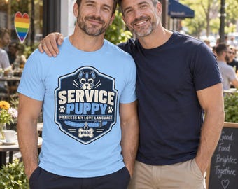 Könnte beinhalten: Ein hellblaues T-Shirt mit einer dunkelblauen schildförmigen Grafik, die ein Welpenmasken-Symbol, den Text "SERVICE PUPPY", "PRAISE IS MY LOVE LANGUAGE" und "WOOF" auf einem knochenförmigen Anhänger zeigt, neben einem dunkelblauen T-Shirt.