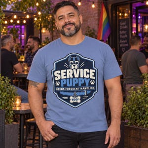 K&ouml;nnte beinhalten: Ein blaues T-Shirt mit einer Grafik eines Hundemasken-Motivs in Schildform, dem Text SERVICE PUPPY, NEEDS FREQUENT HANDLING und einem Hundehalsband mit einer Marke, auf der WOOF steht. Das Design enth&auml;lt Pfotenabdr&uuml;cke und blaue Akzente.