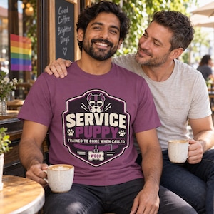 K&ouml;nnte beinhalten: Ein malvenfarbenes T-Shirt mit einer Grafik eines Hundes, der ein Geschirr tr&auml;gt, innerhalb eines schildf&ouml;rmigen Emblems. Der Text lautet SERVICE PUPPY, TRAINED TO COME WHEN CALLED und WOOF auf einem knochenf&ouml;rmigen Anh&auml;nger.