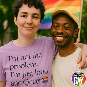 May include: Two people smiling, embracing, and wearing t-shirts with text. One shirt is lavender and says "I'm not the problem. I'm just loud and Queer" with a rainbow flag. The other shirt is cream-colored. A rainbow flag is in the background.