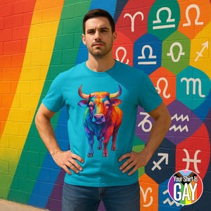 Puede incluir: Una camiseta turquesa con una colorida ilustración de un toro. El toro está representado en tonos vibrantes de rojo, naranja, amarillo, verde y azul. El fondo es una pared de arcoíris con símbolos del zodíaco y el texto "Your Shirt Is GAY".