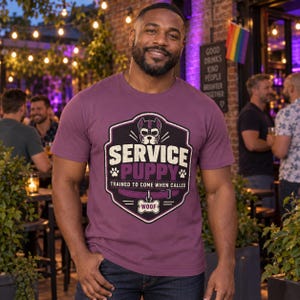 K&ouml;nnte beinhalten: Ein malvenfarbenes Kurzarm-T-Shirt mit einer Grafik eines Hundegesichts in einem Wappen, dem Text SERVICE PUPPY, TRAINED TO COME WHEN CALLED, einem knochenf&ouml;rmigen Anh&auml;nger mit der Aufschrift WOOF, Pfotenabdr&uuml;cken und einem violetten Halsband.