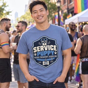 K&ouml;nnte beinhalten: Ein blaues Kurzarm-T-Shirt mit einer Grafik aus Welpenmaske, Halsband und Pfotenabdr&uuml;cken. Der Text lautet SERVICE PUPPY, NEEDS FREQUENT HANDLING und WOOF. Das Design ist in einem dunkelblauen, schildf&ouml;rmigen Emblem mit hellem Rand eingefasst.