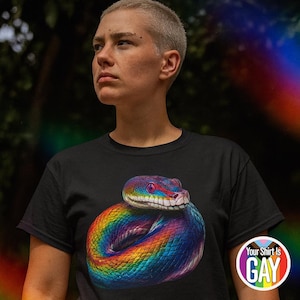 Könnte beinhalten: Schwarzes T-Shirt mit einer Schlange mit Regenbogenfarben. Der Schlangenkopf mit roten Akzenten ist sichtbar. Auf dem T-Shirt befindet sich ein rundes Logo mit der Aufschrift "Your Shirt Is GAY" in einem Regenbogenkreis.