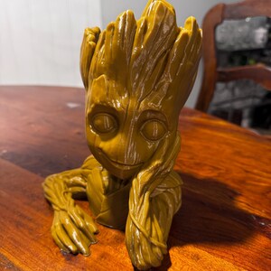 Può includere: Un vaso per piante Groot giallo dorato, con la consistenza del legno e i lineamenti del personaggio. La figura è in posa con il mento appoggiato sulla mano, creando un'espressione pensierosa. Il vaso è progettato per contenere piccole piante o succulente.