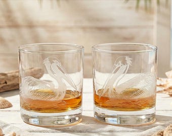 May include: Two clear, round whiskey glasses with etched pelican designs, filled with amber liquid. The glasses sit on a white surface with seashells, a rope, and a linen napkin. The background features a beach scene with palm fronds.