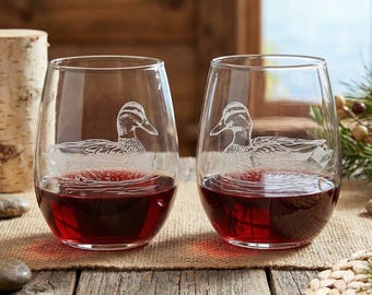 May include: Two stemless wine glasses filled with red wine. Each glass features an etched design of a duck. The glasses are set on a wooden table with natural elements in the background, creating a rustic aesthetic.