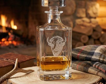 May include: A clear glass decanter with a square base and a stopper, filled with amber liquid. The decanter features an etched bison head design. It sits on a wooden slab with a book, antler, and plaid blanket in the background.