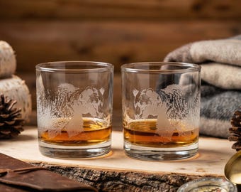 May include: Two clear whiskey glasses with etched bear designs, filled with amber liquid. The glasses sit on a wooden surface, alongside a leather-bound journal, a compass, and pine cones. The background features birch logs and a plaid blanket.