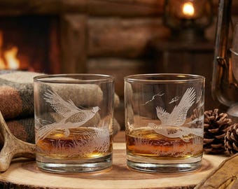 May include: Two clear whiskey glasses with etched pheasant designs, filled with amber liquid. The glasses sit on a wooden slab, with a rustic backdrop of a fireplace, lantern, and gloves. The glasses are approximately 3.5 inches tall.