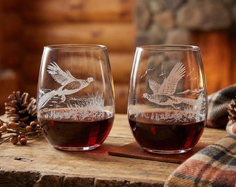 May include: Two stemless wine glasses filled with red wine, each etched with a detailed pheasant design. The glasses sit on a rustic wooden table, with a cozy plaid blanket and a lit fireplace in the background.