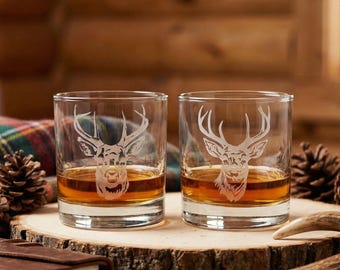 May include: Two clear whiskey glasses with etched deer head designs. Each glass is filled with amber liquid and sits on a wooden slab. The glasses are surrounded by pine cones, a leather-bound journal, and a deer antler.
