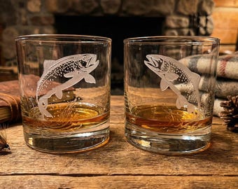 May include: Two clear whiskey glasses with etched trout designs, filled with amber liquid. The glasses sit on a wooden surface, alongside a fishing fly and a leather-bound journal. The background includes a stone fireplace and a cozy blanket.