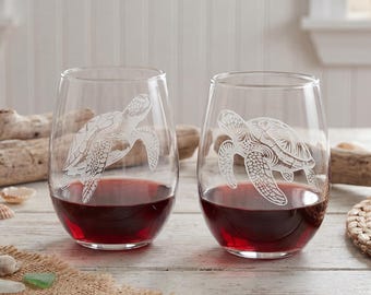 May include: Two stemless wine glasses filled with red wine. Each glass features an etched design of a sea turtle. The glasses are on a light-colored wooden surface, with beach-themed decorations.