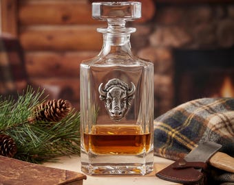 May include: A clear glass decanter with a silver bison head emblem, filled with amber liquid. The decanter has a square shape and a rectangular stopper. The image is set against a rustic background with a plaid blanket and an axe.