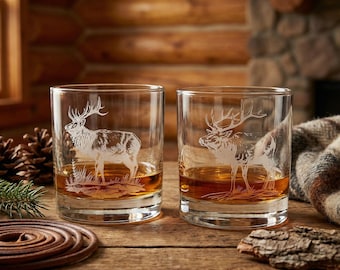 May include: Two clear whiskey glasses with etched elk designs. The glasses are filled with amber liquid and sit on a wooden surface. The background features a rustic log cabin aesthetic, with a fireplace and wooden walls.
