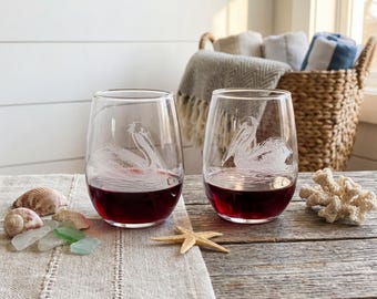 May include: Two stemless wine glasses filled with red wine, each etched with a pelican design. The glasses sit on a linen cloth with seashells, sea glass, and a starfish. A wicker basket with towels is in the background.