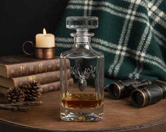 May include: A clear glass decanter with a silver stag head design, filled with amber liquid. The decanter sits on a wooden surface with a lit candle, books, and binoculars in the background. A plaid blanket adds a rustic touch.