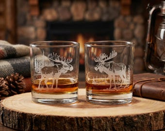 May include: Two clear whiskey glasses with etched moose designs, filled with amber liquid, sit on a wooden slab. The glasses are illuminated, showcasing the moose and forest details. The background includes a fireplace and a lantern.