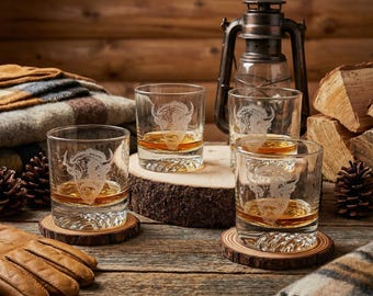 May include: Four clear whiskey glasses with a bison design, filled with amber liquid, sit on wooden coasters. The glasses are arranged on a wooden surface with a rustic lantern and firewood in the background. A pair of leather gloves and a plaid blanket are also visible.