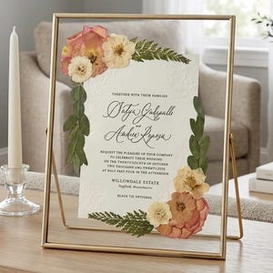 May include: A rectangular gold metal floating frame containing a white wedding invitation with embossed floral details. The invitation is decorated with pressed pink and cream flowers and green fern leaves, featuring black calligraphy text.