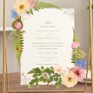May include: A rectangular gold metal frame containing a wedding invitation decorated with pressed flowers and green ferns. The invitation text includes the phrase Together with their families, request the pleasure of your company at the celebration of their marriage, August 8, 2026 at 3 PM, and Dinner reception to follow.