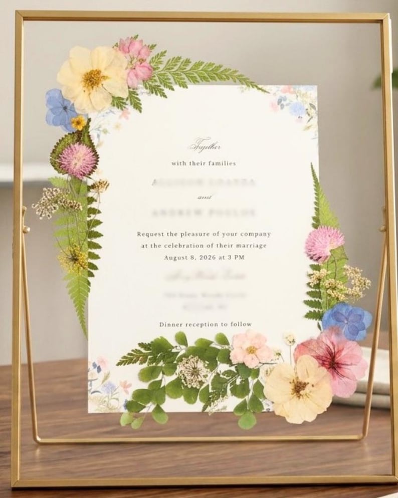May include: A rectangular gold metal frame containing a wedding invitation decorated with pressed flowers and green ferns. The invitation text includes the phrase Together with their families, request the pleasure of your company at the celebration of their marriage, August 8, 2026 at 3 PM, and Dinner reception to follow.