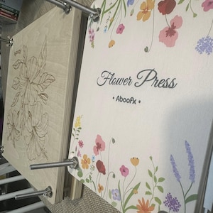 A wooden flower press with four metal bolts and wing nuts. This will help with edding flower preservation and also create a beautiful piece of art. The top board features a colorful floral print with the text "Flower Press" and "Aboofx". The bottom board displays a laser-engraved hibiscus flower design.