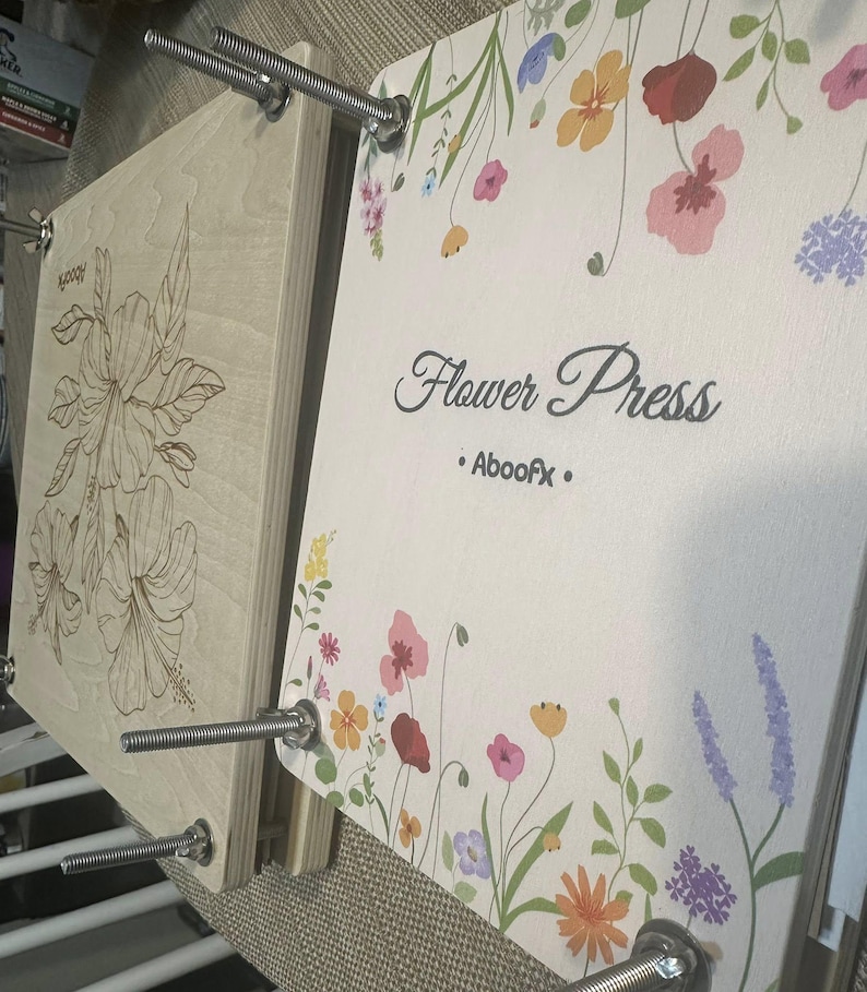 A wooden flower press with four metal bolts and wing nuts. This will help with edding flower preservation and also create a beautiful piece of art. The top board features a colorful floral print with the text "Flower Press" and "Aboofx". The bottom board displays a laser-engraved hibiscus flower design.