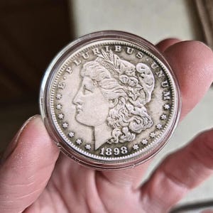 May include: A silver coin from 1898, featuring a profile of a woman and the inscription "E PLURIBUS UNUM." The coin is encased in a clear plastic holder, held by a hand. The coin's edge is detailed with stars.