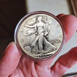 Peut inclure: Une pièce de monnaie en argent, un demi-dollar Walking Liberty daté de 1943, dans un étui de protection transparent. La pièce représente Lady Liberty marchant vers le soleil, avec l'inscription "IN GOD WE TRUST".