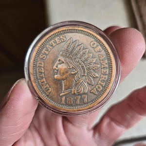 Peut inclure: Une pièce de monnaie américaine Indian Head de 1877, de couleur cuivrée, dans un support de protection transparent. La pièce représente un profil d'Amérindien coiffé d'une coiffe de plumes et les mots "LIBERTY" et "UNITED STATES OF AMERICA".