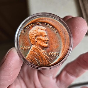 Może przedstawiać: Miedziowy grosz Lincolna z 1955 roku w przezroczystym etui ochronnym. Moneta przedstawia profil Abrahama Lincolna oraz napisy "IN GOD WE TRUST" i "LIBERTY". Widoczny jest również rok "1955".