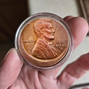May include: A copper 1946 Lincoln penny in a clear plastic case. The coin shows a profile of Abraham Lincoln, with the word "LIBERTY" and the year "1946" visible. The coin is held in a person's hand.