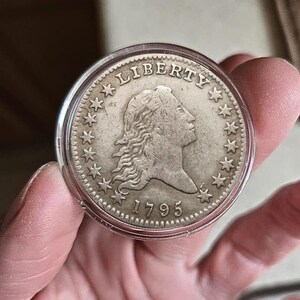 Peut inclure: Une pièce de monnaie en argent de 1795, représentant un profil de femme et le mot "LIBERTY" en haut. La pièce est entourée d'étoiles et est enfermée dans un support en plastique transparent.