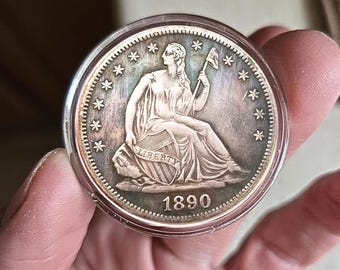 Może przedstawiać: Srebrna moneta z siedzącą postacią trzymającą flagę i tarczę, z napisem "LIBERTY" i rokiem 1890. Moneta jest w przezroczystym etui ochronnym, trzymana w dłoni.