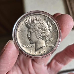 May include: A silver coin from 1921, featuring a profile of a woman with rays emanating from her head. The coin is encased in a clear protective holder. Inscriptions include "IN GOD WE TRUST" and "LIBERTY".
