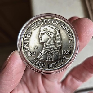 Peut inclure: Une pièce de monnaie Half Dollar en argent de 1921, dans un étui de protection transparent. La pièce représente un profil de femme et les inscriptions "UNITED STATES OF AMERICA" et "HALF DOLLAR". Les années 1821 et 1921 sont également visibles.