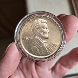 Puede incluir: Una moneda de un centavo Lincoln de 1955 en un estuche protector. La moneda presenta un perfil de Abraham Lincoln y las palabras "IN GOD WE TRUST" y "LIBERTY". El año "1955" es visible. La moneda está en un soporte de plástico transparente.