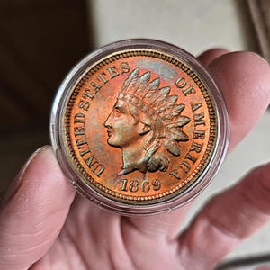 Peut inclure: Une pièce de monnaie Indian Head de 1869, dans un étui de protection transparent. La pièce en cuivre représente un profil d'Amérindien avec une coiffe de plumes et les mots "UNITED STATES OF AMERICA". La pièce est de couleur brun-rouge.